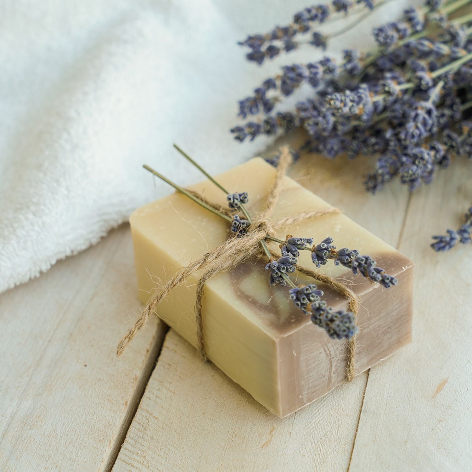 Savon artisanal lavande-patchouli sur su bois, emballé dans une corde de jute avec des petites branches de lavande sur le dessus, comme un cadeau. À l'arrière, une serviette blanche et d'autres branches de lavande.